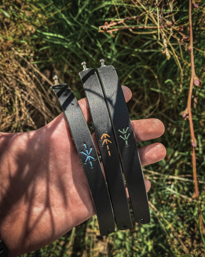 Set of three handcrafted black leather Viking bracelets, each stitched with a unique Norse rune representing victory, protection, and hospitality in tan, green, and blue thread.